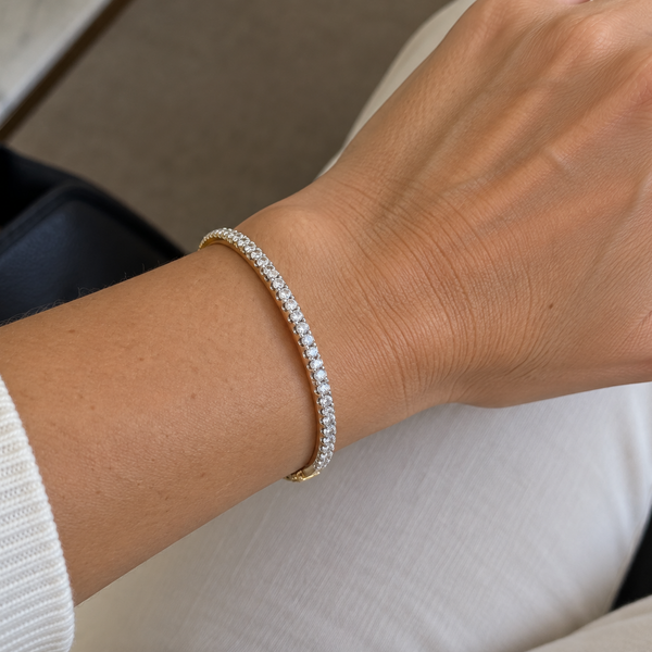 A close-up of a person's wrist wearing a delicate tennis bracelet with small clear stones set in a yellow gold band. The person is wearing a white ribbed sweater and white pants, with a black handbag visible in the background.