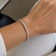 A close-up of a person's wrist wearing a delicate tennis bracelet with small clear stones set in a yellow gold band. The person is wearing a white ribbed sweater and white pants, with a black handbag visible in the background.